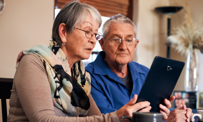 An elderly couple sits together at a dining table using a tablet, demonstrating senior independence, digital engagement, and supportive in-home care that helps older adults stay connected and informed in their own home.